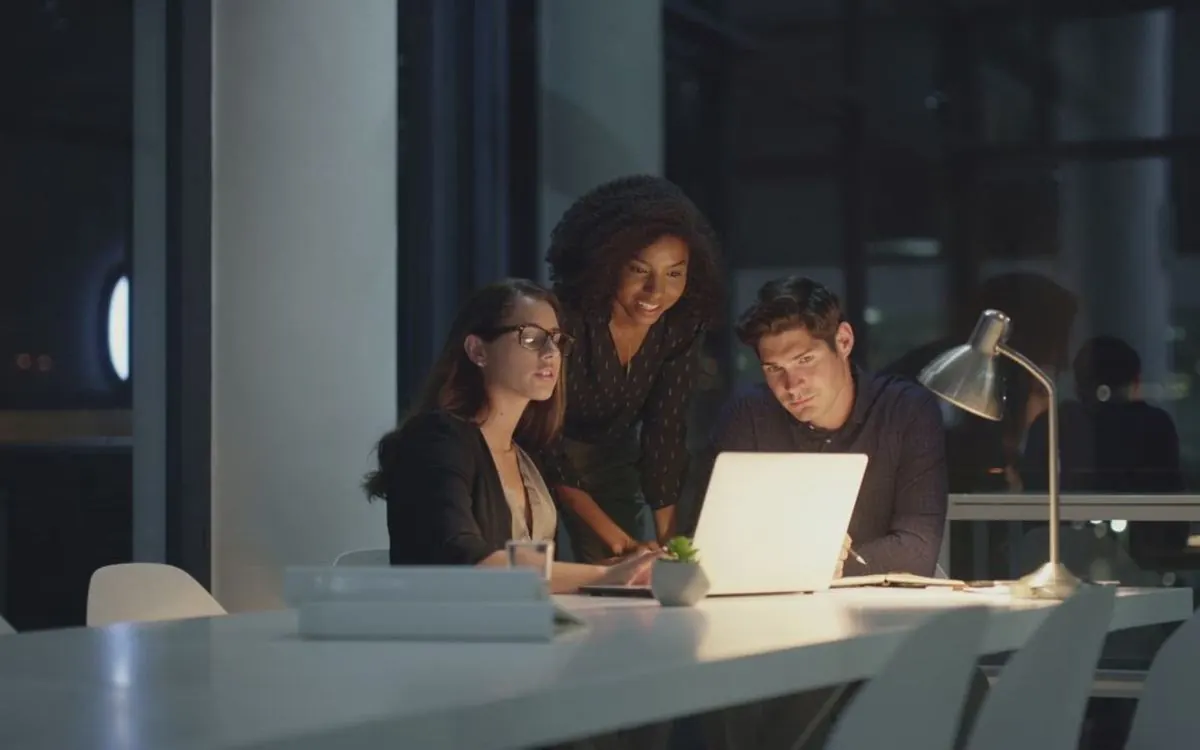 Três profissionais reunidos em volta de um notebook à noite